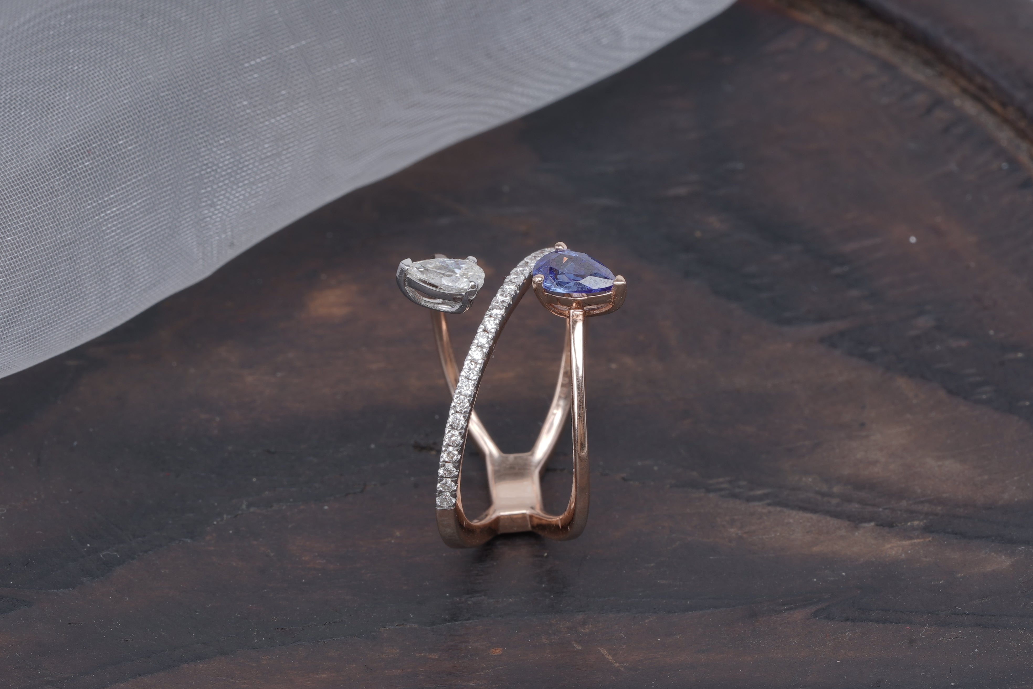 Modern 18K rose gold crossover ring with pear-shaped sapphire, diamond, and pavé band